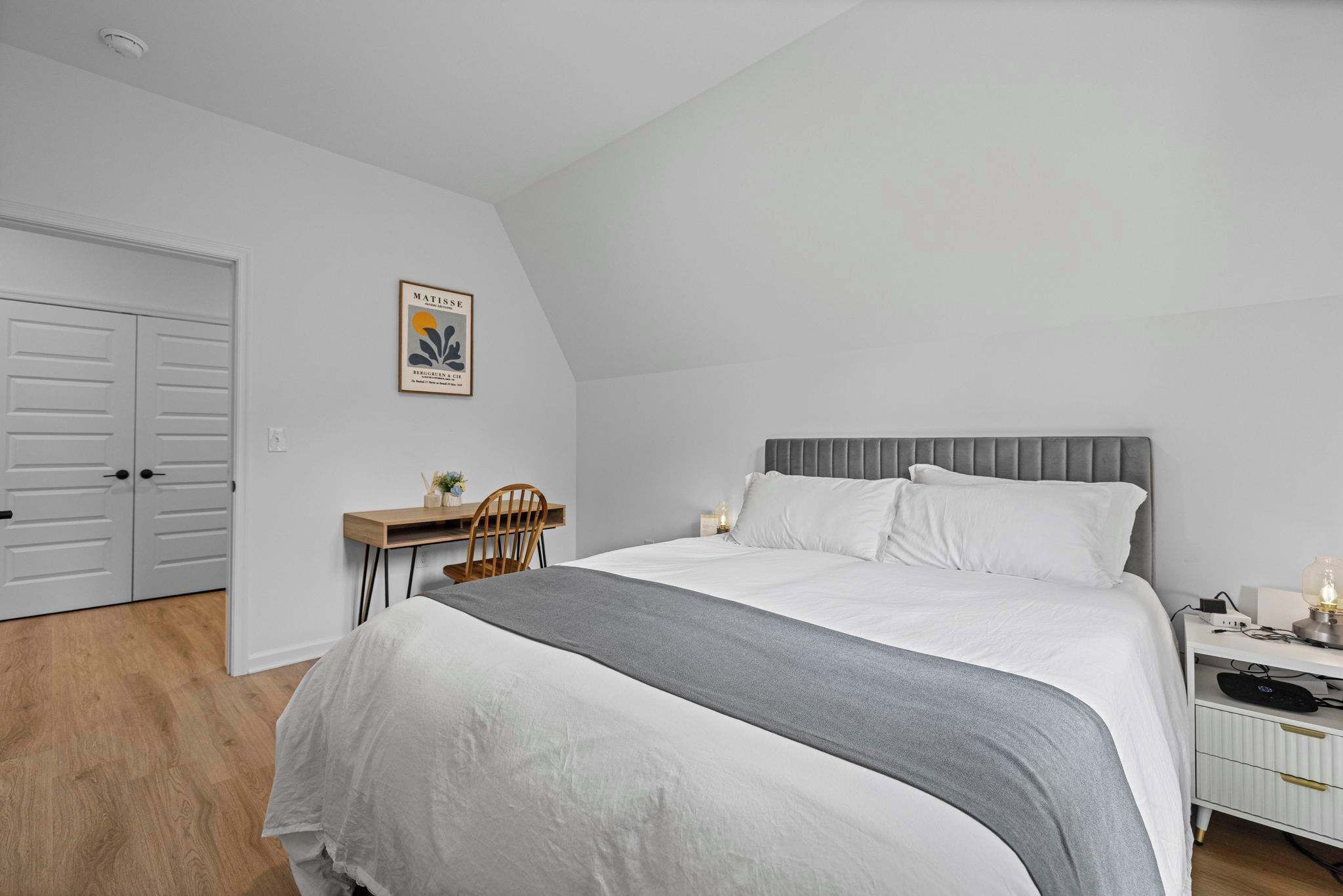 A bedroom with a bed, bedside table, chair, desk, and a framed picture on the wall.