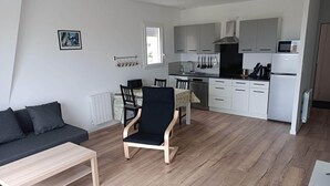 Cocina moderna con armarios blancos, un microondas y un lavavajillas. Hay un comedor con mesa y sillas, y un sofá negro en la sala de estar.