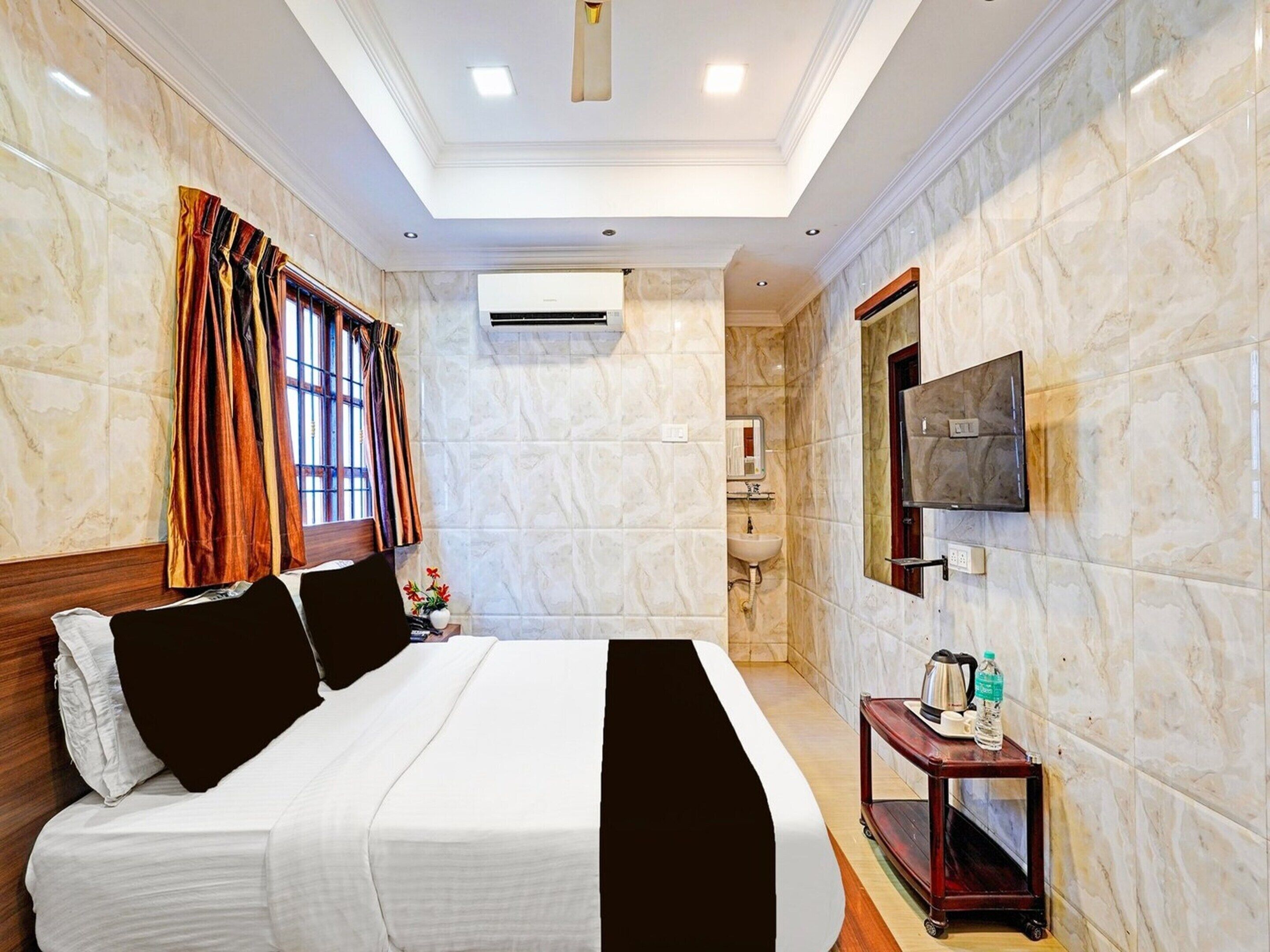 A hotel room with a bed, a TV mounted on the wall, a bedside table with a kettle, and a window with curtains.