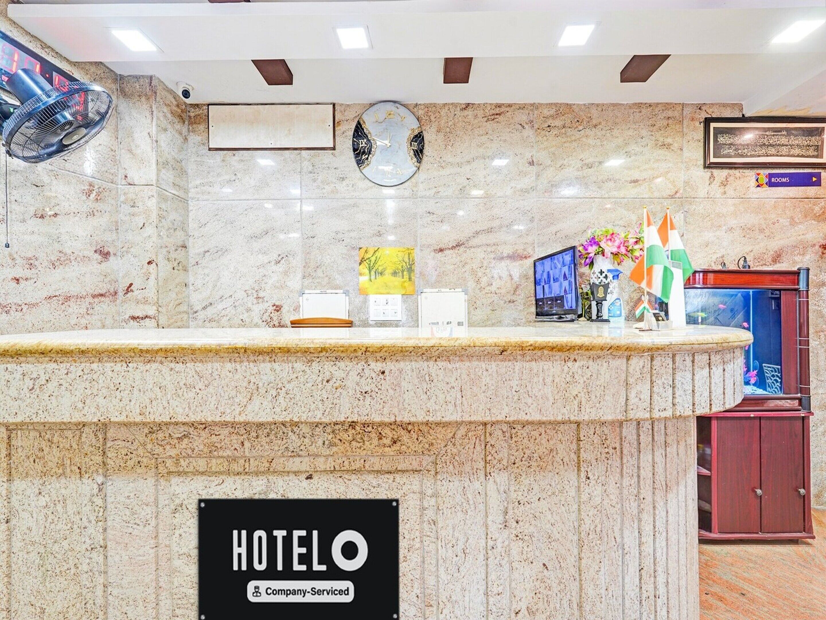 A hotel reception desk with a clock, a digital display, a sign indicating it's company-serviced, and a sign for rooms.