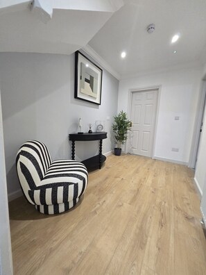 Ein Zimmer mit Holzboden, einem schwarz-weiß gestreiften Stuhl, einem kleinen schwarzen Tisch mit Dekoration und einem gerahmten Bild an der Wand, sowie einer weißen Tür.