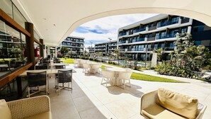 A modern outdoor patio with wicker furniture, a pool, and a view of multi-story buildings. - Makia Beach Resort Luxo - Muro Alto-10 Piscinas (Ipojuca)