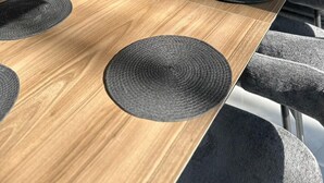 Un tavolo da pranzo moderno con piano in legno, sottopiatti circolari neri e un vaso con piante essiccate.