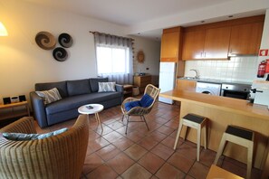 Un espace de vie compact comprenant un canapé, deux fauteuils en osier, une petite table ronde, un coin cuisine avec des meubles en bois, un lave-linge et un sol carrelé.