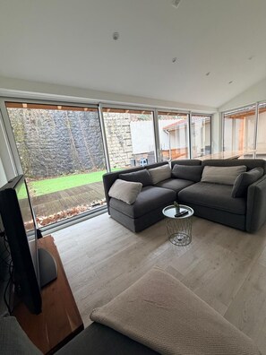 Una sala de estar moderna con un sofá modular, una pequeña mesa redonda y un televisor sobre un mueble de madera. Amplias ventanas ofrecen vistas a un muro de piedra y un patio cubierto de césped.