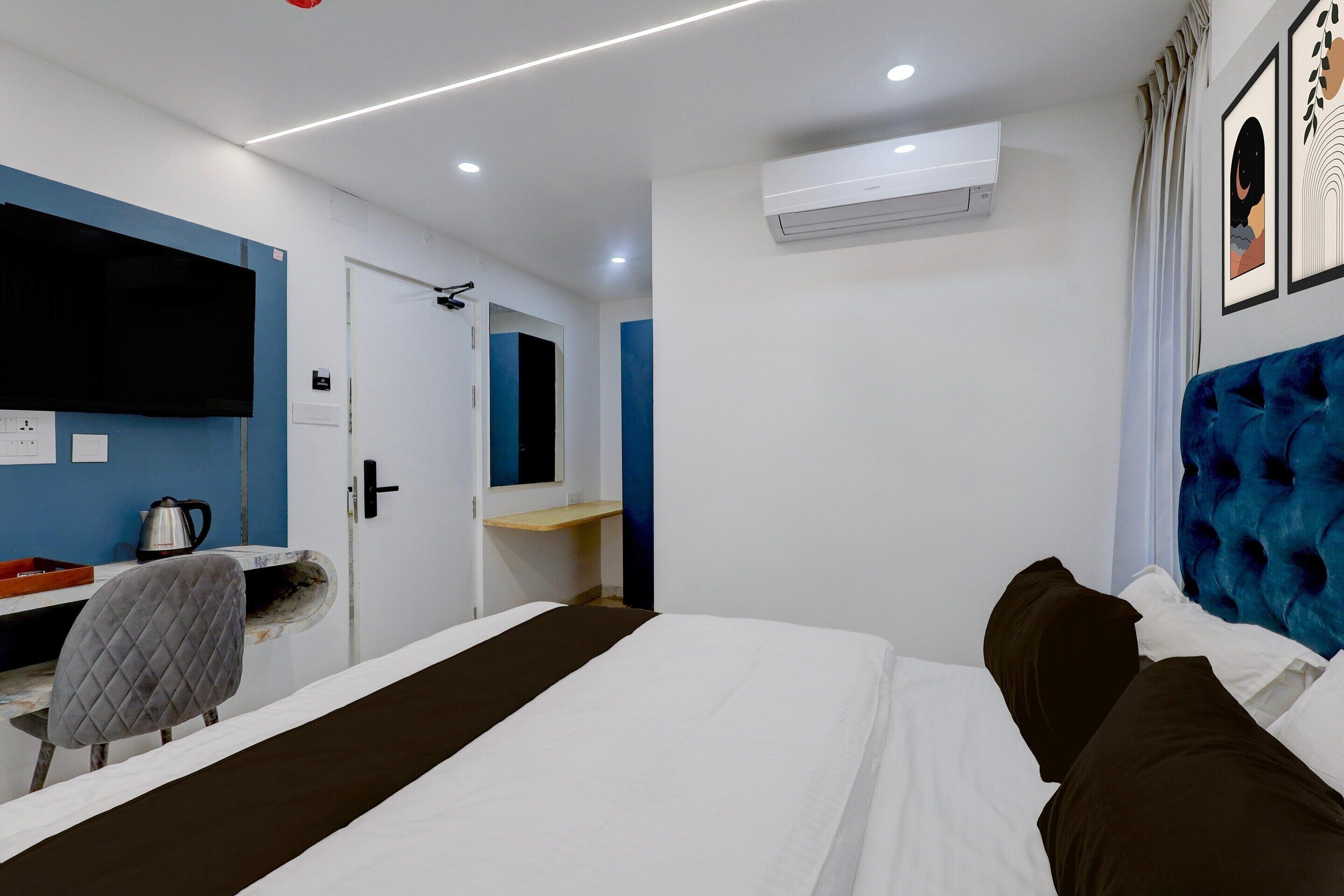 A hotel room with a bed, a desk with a chair, a flat-screen TV, an air conditioning unit, and a blue headboard.