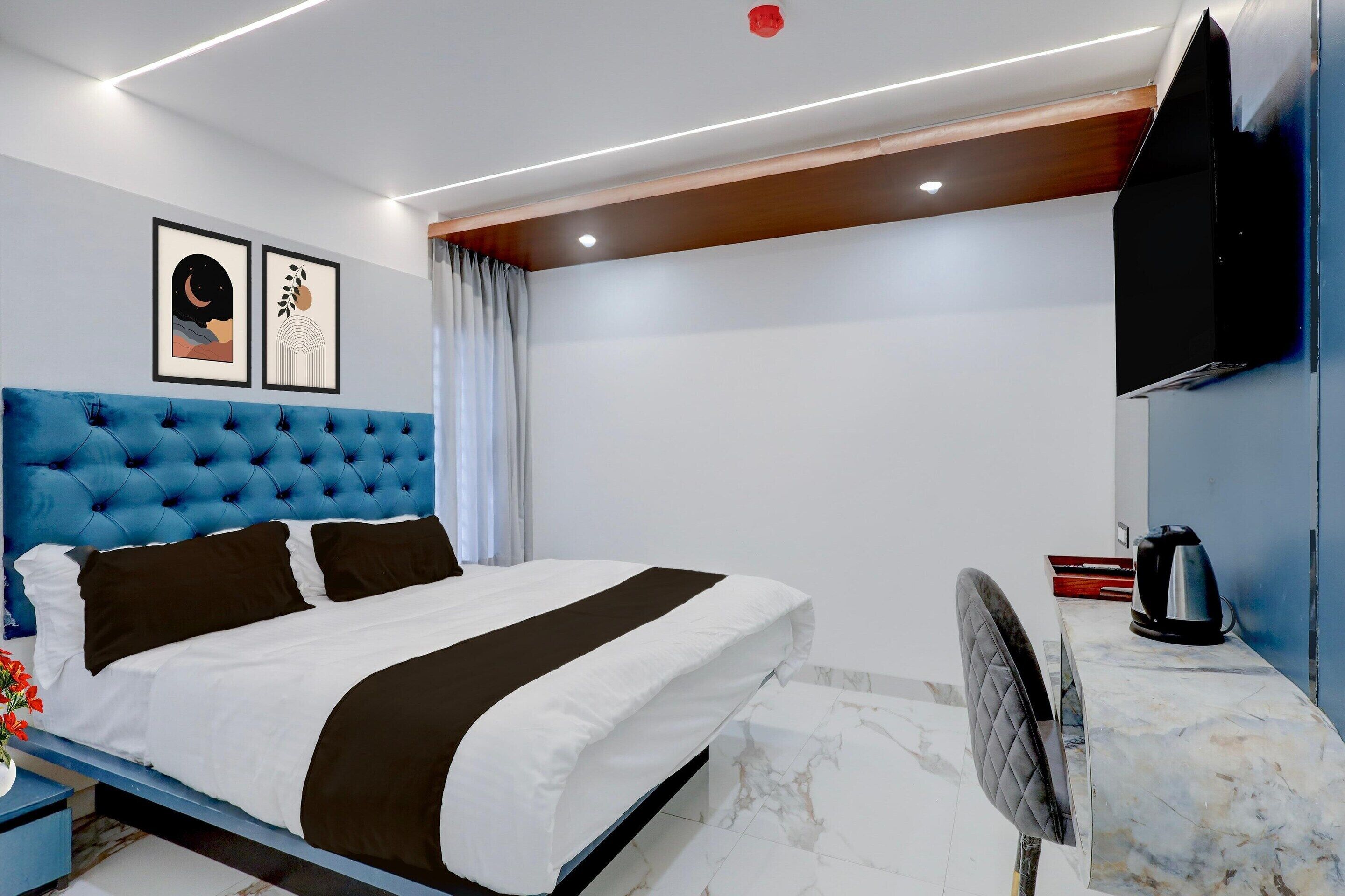 A modern hotel room with a tufted headboard, a bed with white and brown bedding, a flat-screen TV mounted on the wall, a marble-tiled floor, and a small table with a kettle.