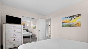 Une chambre à coucher avec un lit, une commode, une porte donnant sur une autre pièce et un tableau accroché au mur.