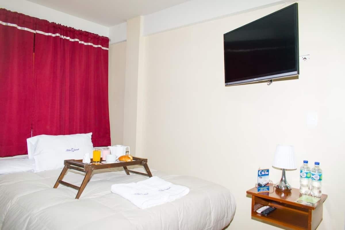 A hotel room with a bed, a TV, a tray with breakfast items, a bedside table with a lamp and water bottles, and red curtains.