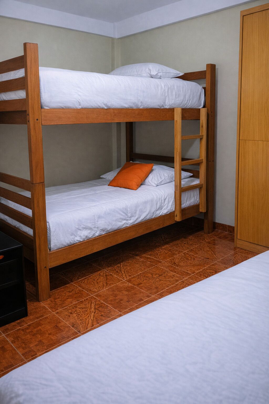 A bunk bed with white bedding and an orange pillow, wooden headboard, and a wardrobe in a room with tiled flooring.