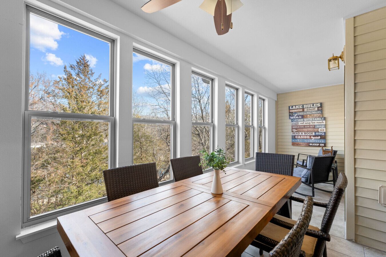A sunroom with large windows, a wooden table, wicker chairs, and a poster on the wall.