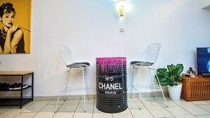 Ein Zimmer mit einem schwarzen Behälter, beschriftet mit 'No 5 Chanel Paris', zwei weißen Drahtstühlen, einem kleinen Tisch mit Schuhen, einer Topfpflanze und einem Fernseher.