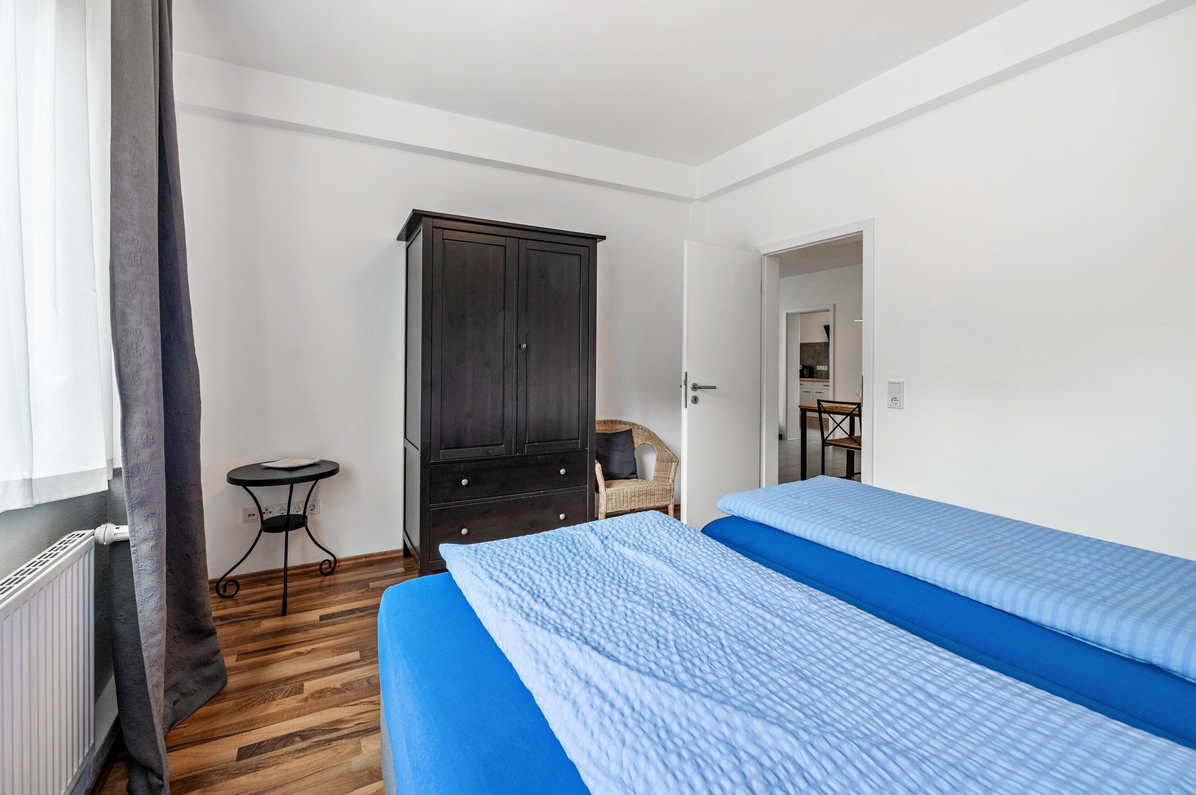 A bedroom with two beds, a dark wooden wardrobe, a small table, and a wicker chair.