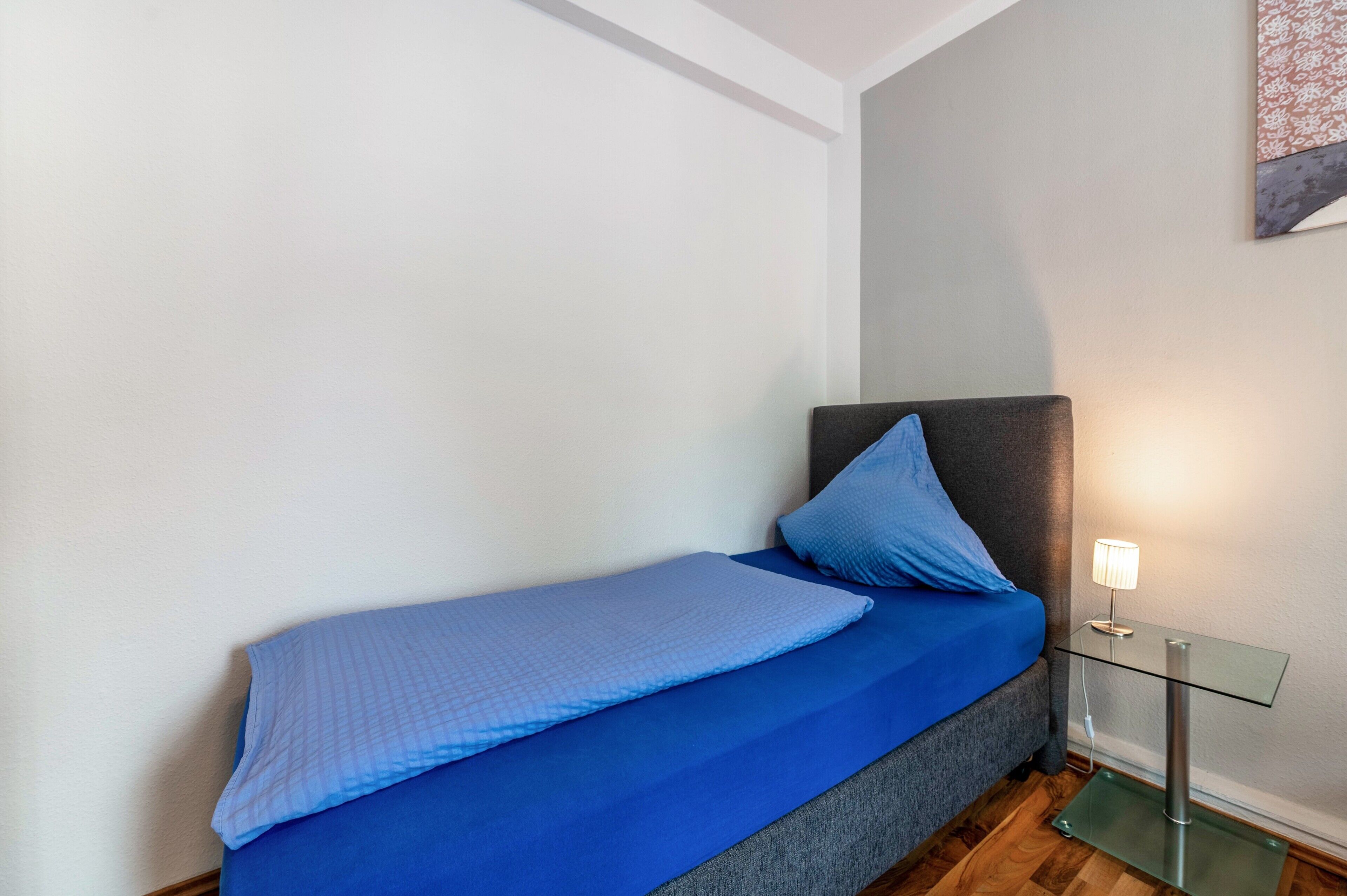Un letto singolo con copriletto e cuscino blu, un comodino in vetro con una lampada e una testiera grigia.