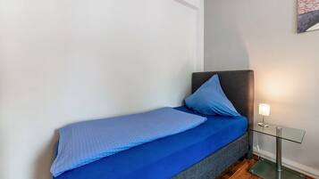 Un letto singolo con copriletto e cuscino blu, un comodino in vetro con una lampada e una testiera grigia.