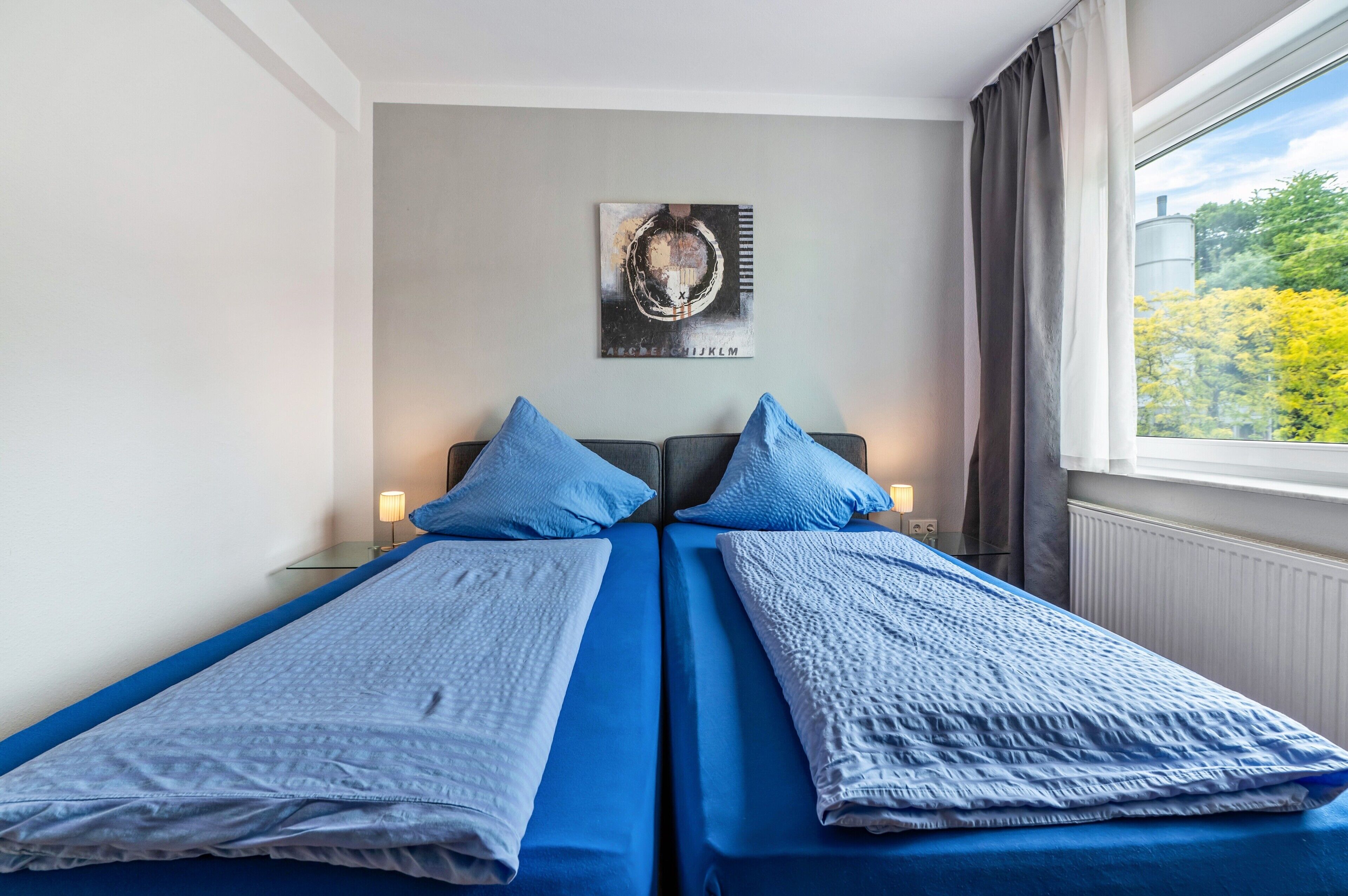 Una cama doble con ropa de cama azul, un cuadro colgado en la pared y una ventana con vistas a árboles.