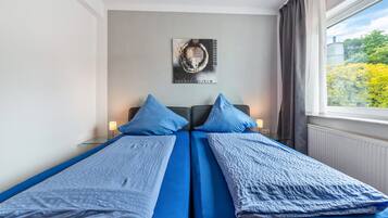 Una cama doble con ropa de cama azul, un cuadro colgado en la pared y una ventana con vistas a árboles.