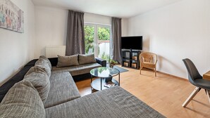 Un salon moderne avec un canapé d’angle, une table basse en verre, un téléviseur à écran plat et un fauteuil en osier.