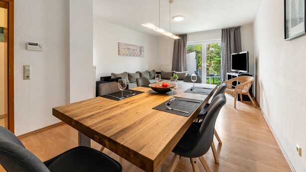 Un salon moderne avec une table à manger en bois, des chaises noires, un ensemble de canapés, un téléviseur et une grande fenêtre avec des rideaux.