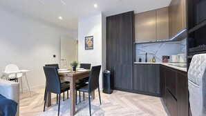 Eine moderne Küche mit dunklen Schränken, ein Essbereich mit Holztisch und -stühlen, sowie ein kleiner weißer Stuhl.