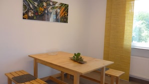 Un petit coin repas avec une table en bois, deux bancs, une petite plante en pot et une fenêtre avec des stores jaunes.