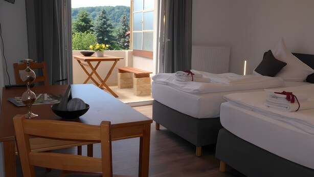 Une chambre d’hôtel avec un lit, une table à manger avec des chaises, une petite table avec un vase et des fleurs, et une vue sur la verdure à l’extérieur.