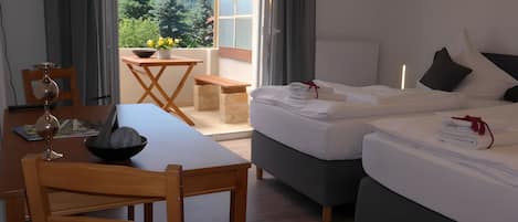 A hotel room with a bed, a dining table with chairs, a small table with a bowl and flowers, and a view of greenery outside.