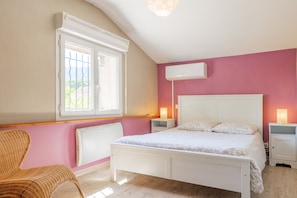 Une chambre avec un lit blanc, deux tables de chevet, une chaise en osier, une fenêtre avec des stores et un appareil de climatisation.