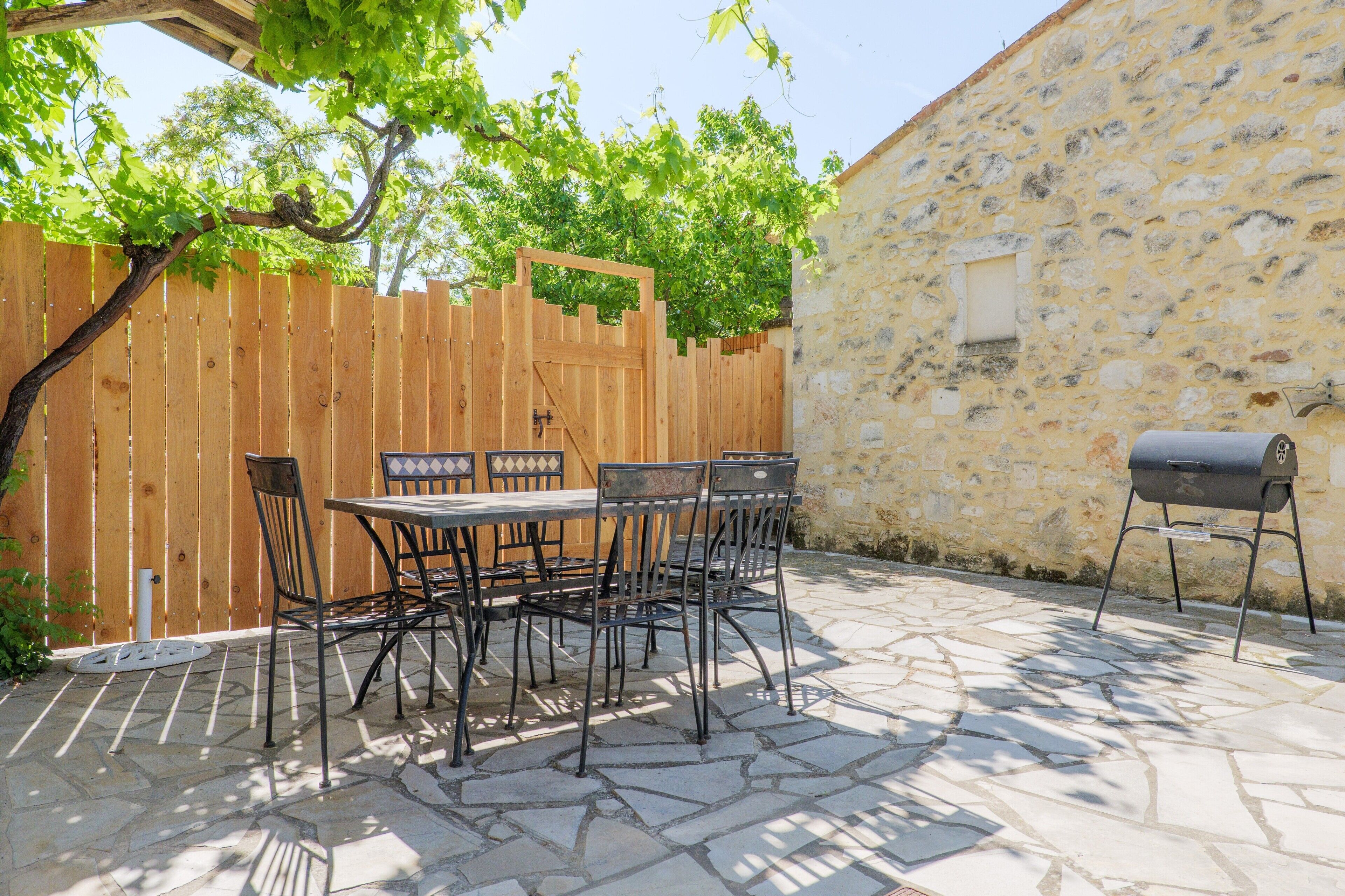 Une terrasse avec un sol en pierre, une clôture en bois, une table avec des chaises et un barbecue.