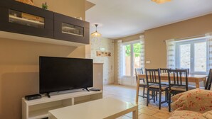 A living room with a TV, white furniture, a dining area with chairs, and a window with curtains.