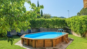 A rectangular wooden above-ground pool with a ladder, surrounded by a grassy yard and a hedge.
