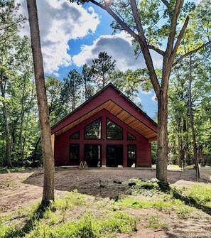 A modern cabin with a red exterior, large windows, and a sloped roof, surrounded by a forest.