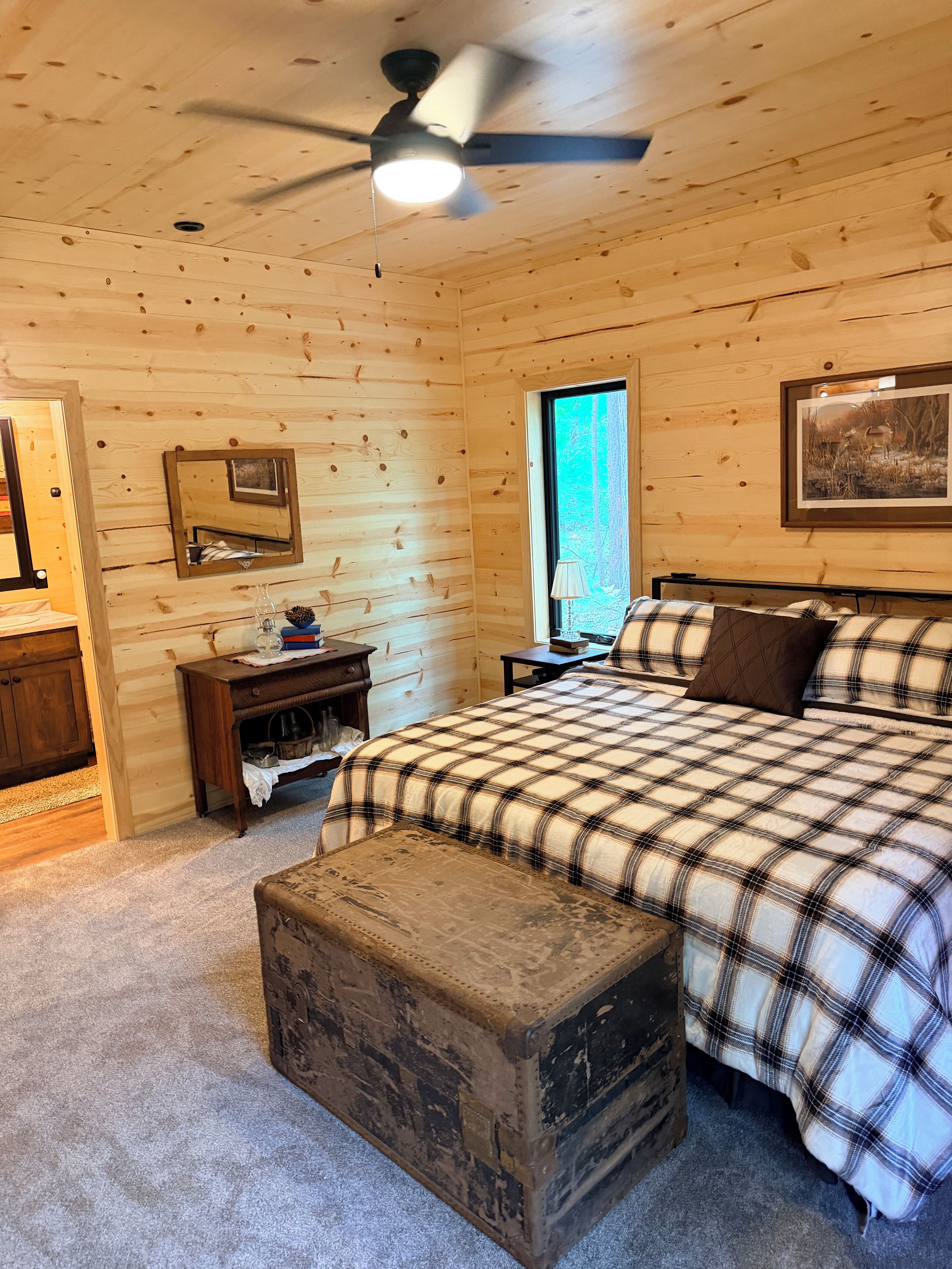 Quarto de cabana de madeira com cama, mesa de cabeceira, cômoda, ventilador de teto e janela.