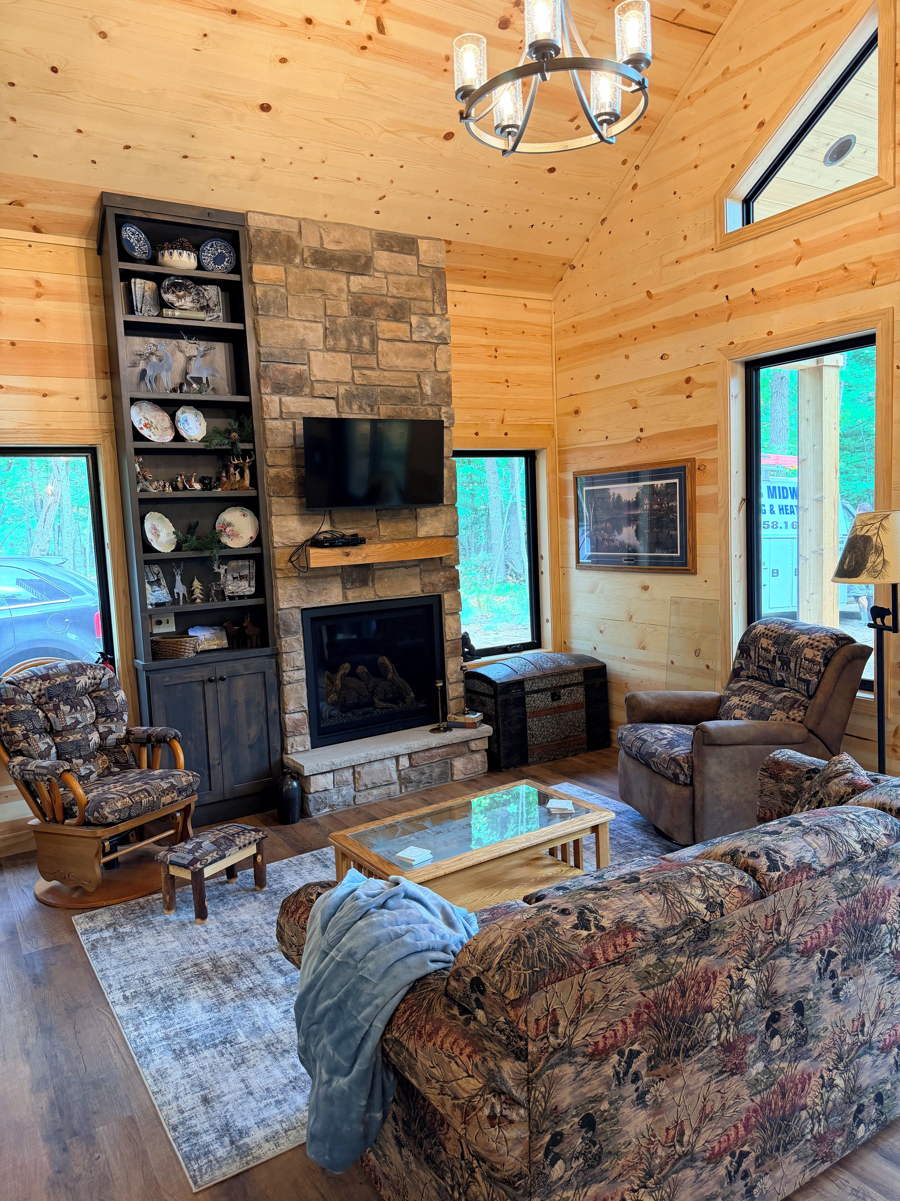 A cozy cabin interior with a stone fireplace, wooden walls, a television, and a large window.