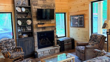 A cozy cabin interior with a stone fireplace, wooden walls, a television, and a large window.