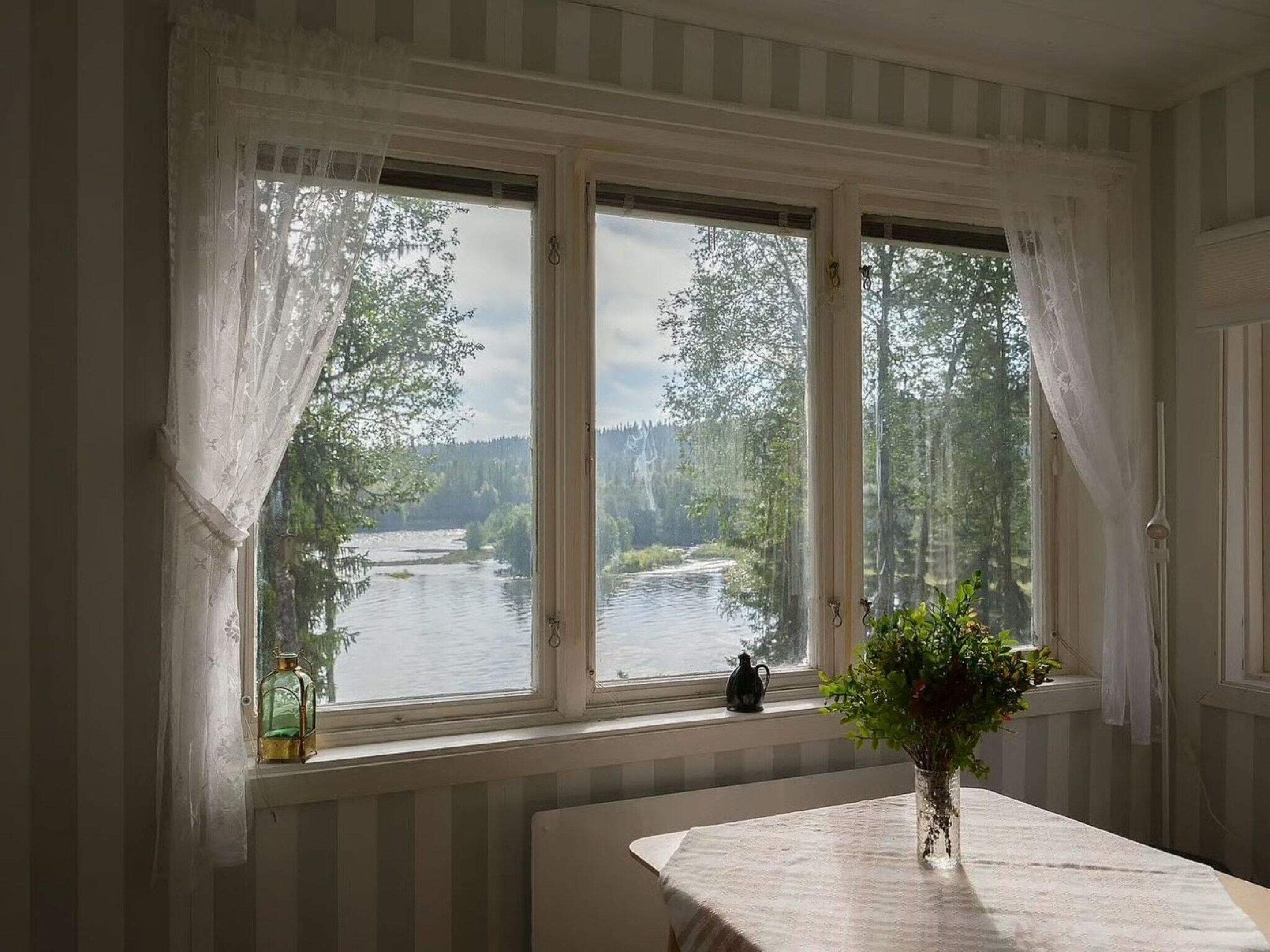 Una habitación con un ventanal que da a un río y un bosque, una mesa con un florero y un farol en el alféizar.