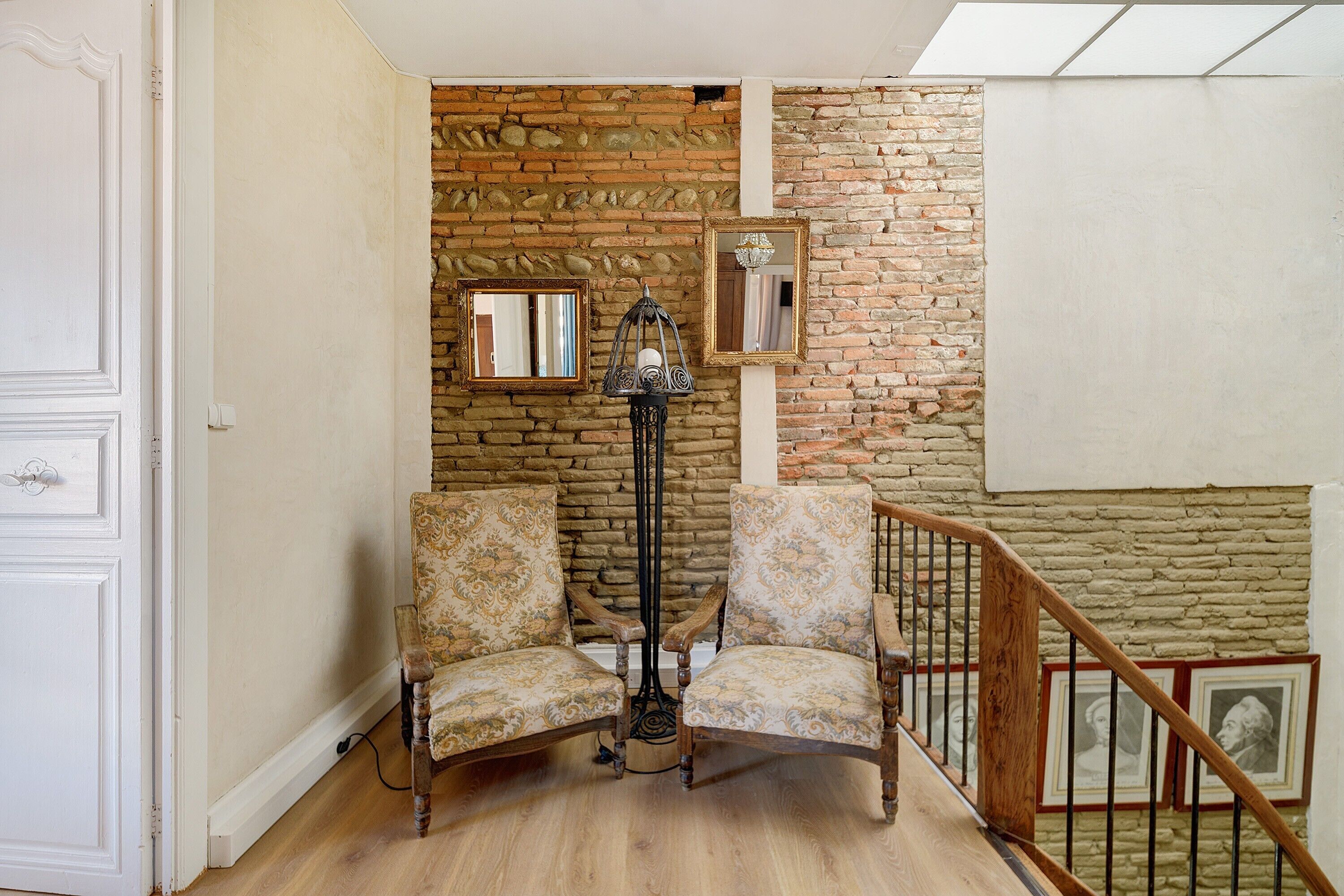 A room with a brick wall, two patterned chairs, a floor lamp, and a staircase.