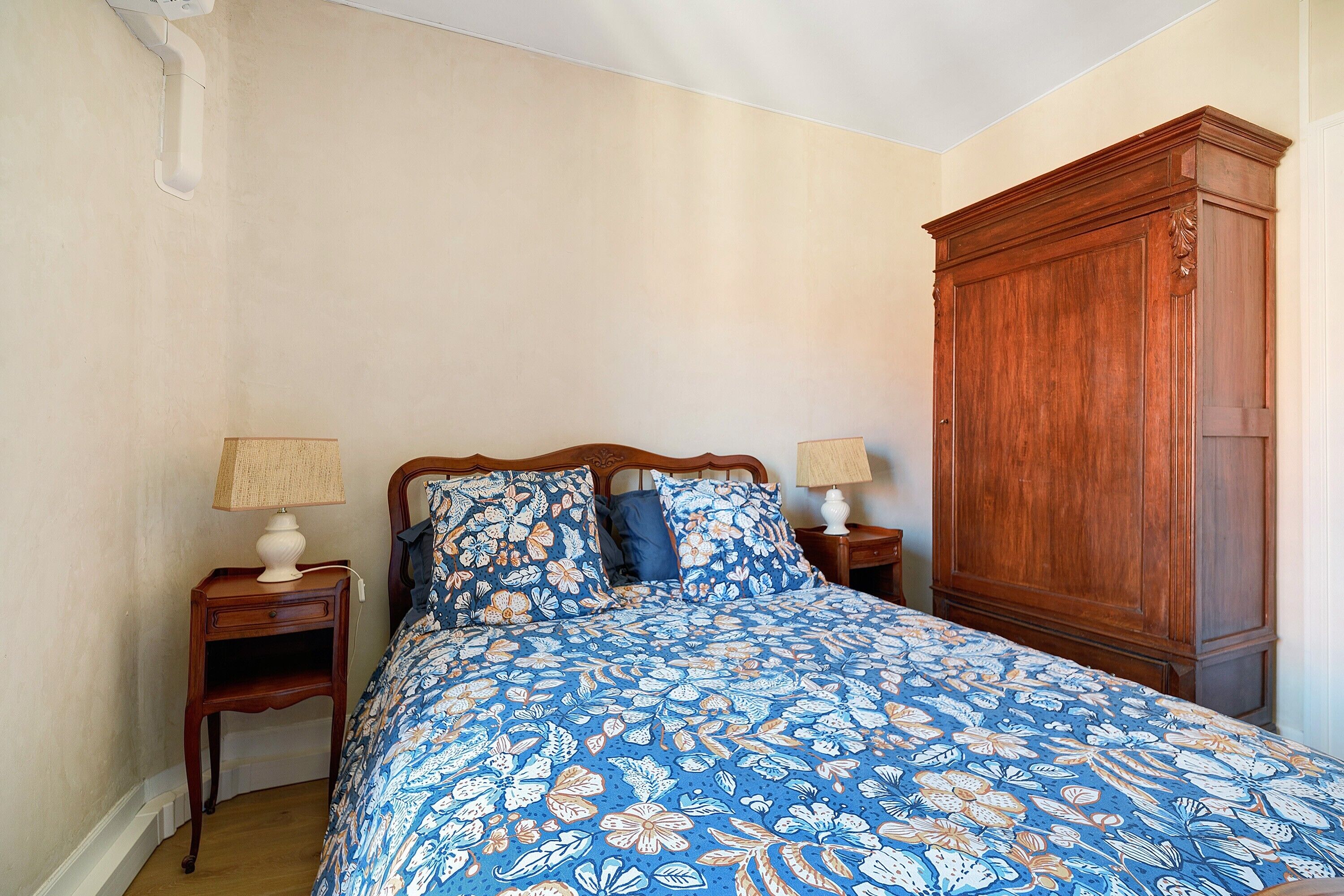 Een slaapkamer met een bed, twee nachtkastjes, een houten garderobe en een bloemenprint beddeken.