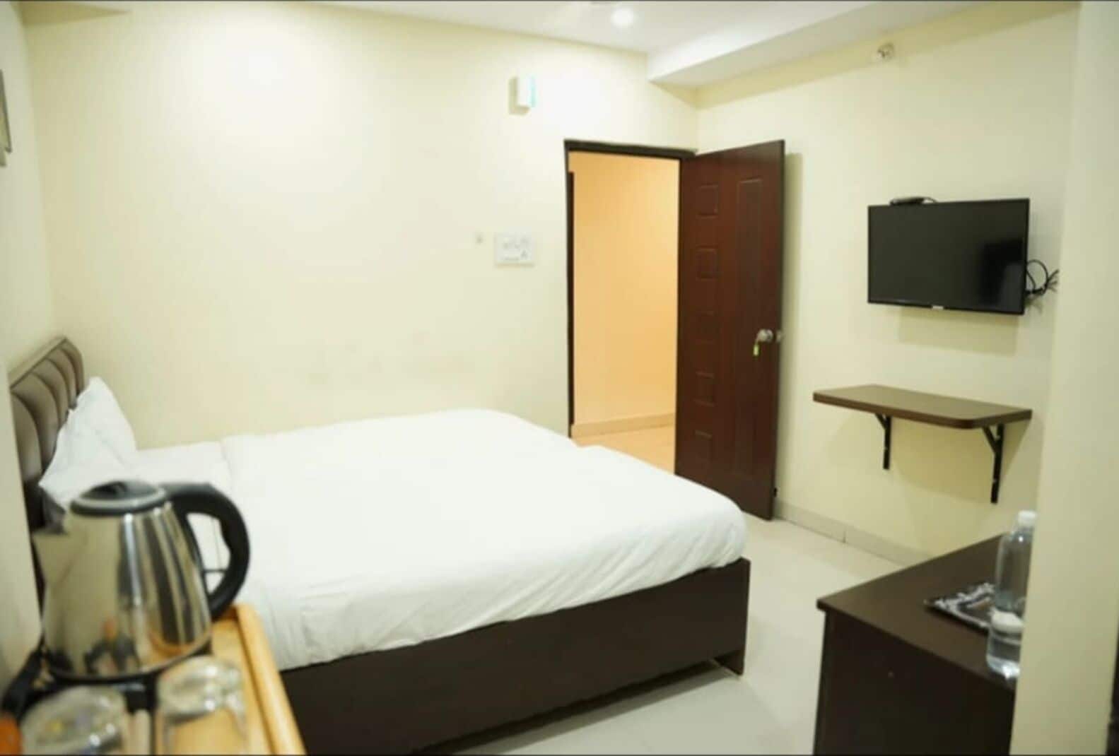 Une chambre d’hôtel avec un lit, un téléviseur fixé au mur, une petite table avec une bouilloire et une porte donnant sur une autre pièce.