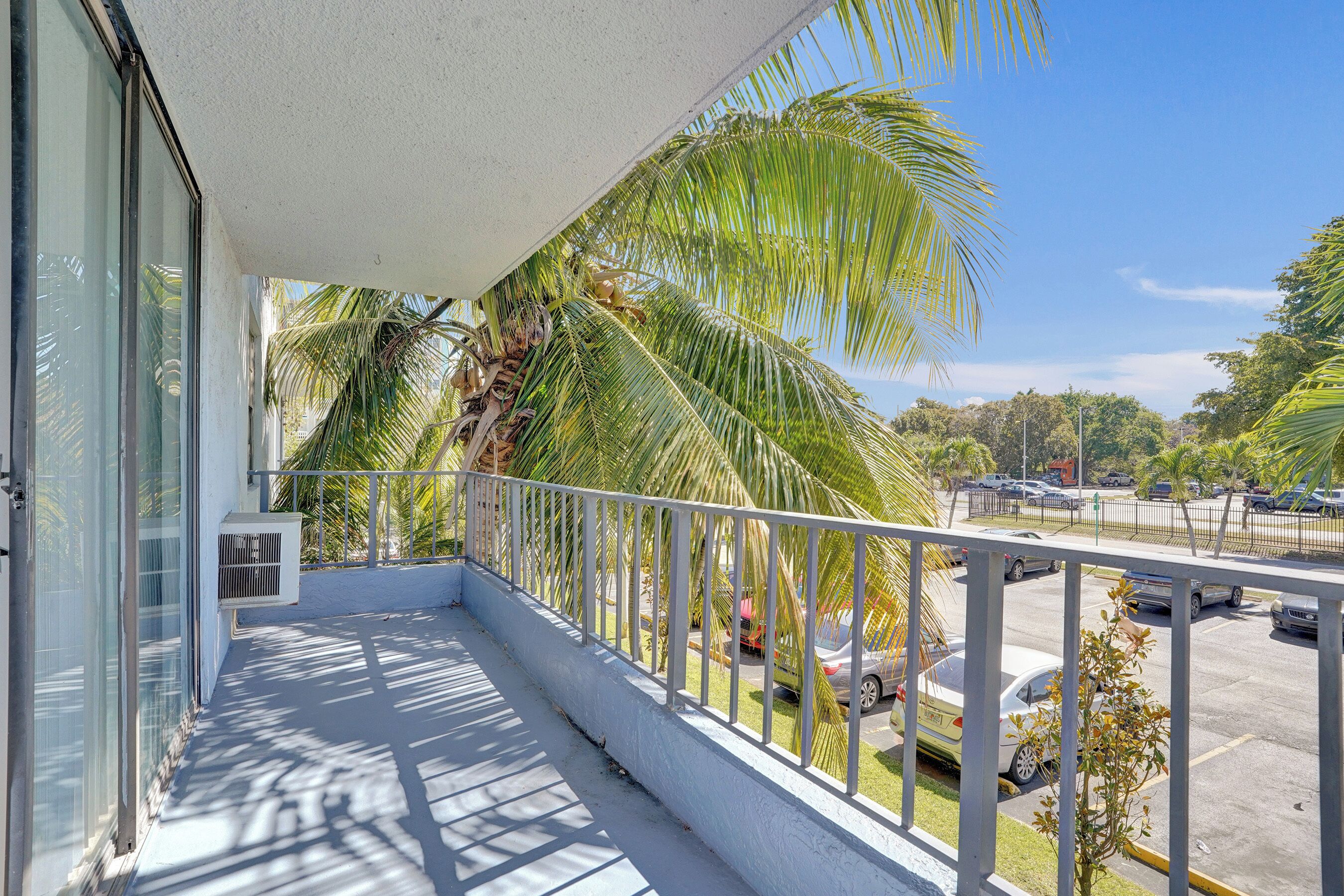 A balcony with a white railing, a glass door, and a view of parked cars and palm trees.