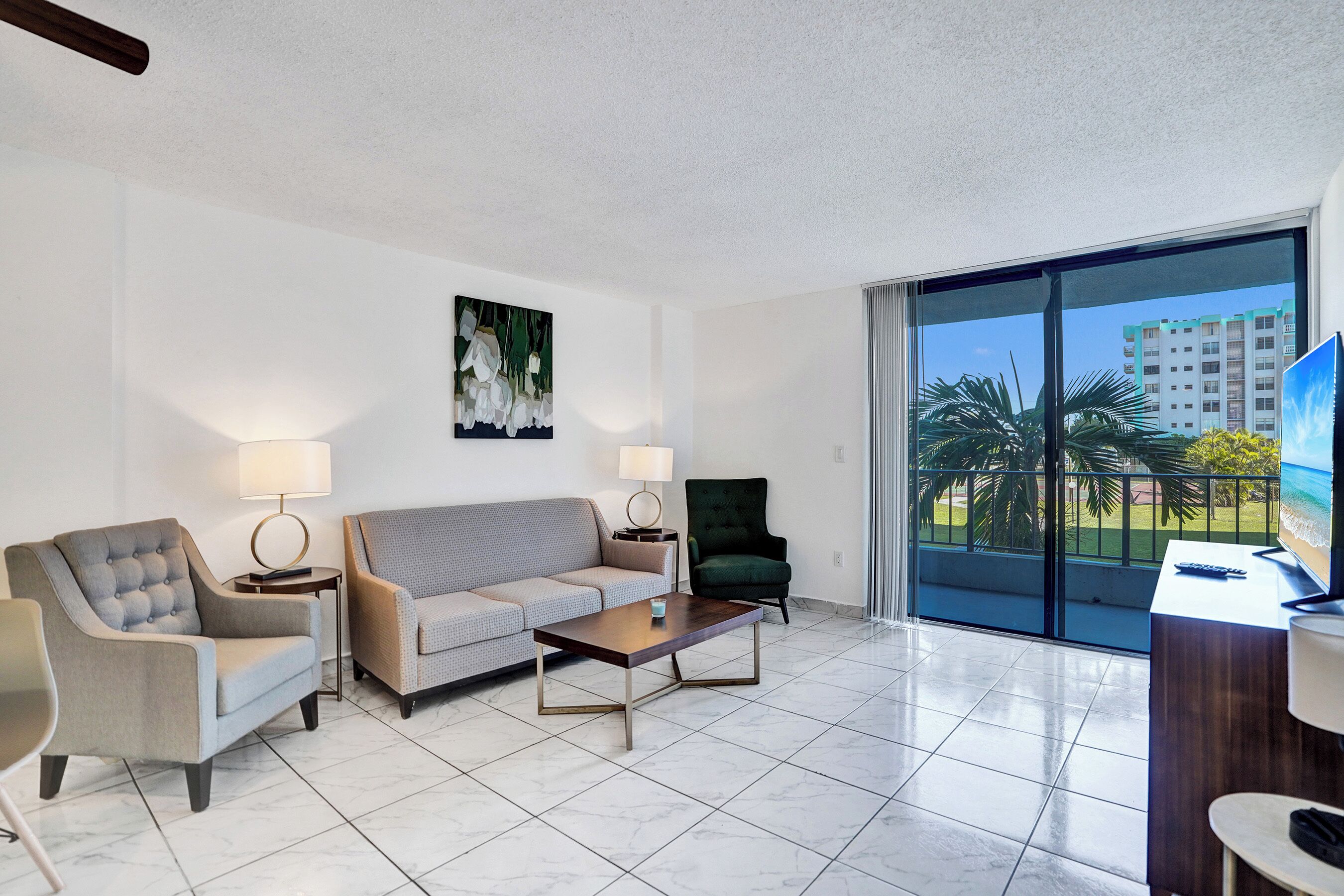 A hotel room with a sofa, armchairs, a coffee table, a TV, and a balcony with a view of palm trees and buildings.
