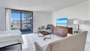 Ett hotellrum med en säng, en soffa, en tv, ett skåp, ett skrivbord och ett stort fönster med utsikt över byggnader.