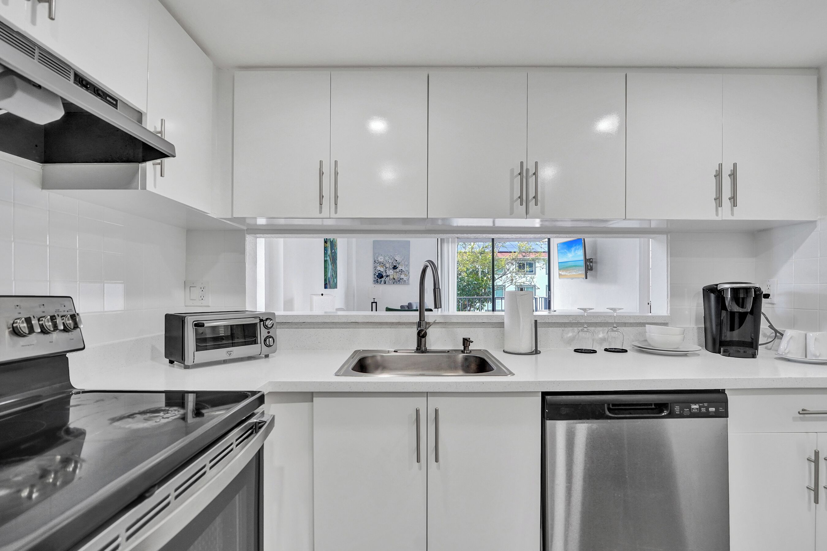 A modern kitchen with white cabinets, a stainless steel dishwasher, a built-in oven, and a window with a view of the outdoors.