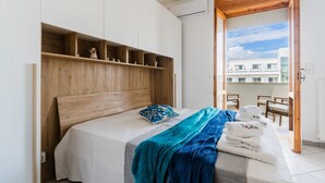 A bedroom with a wooden headboard, a bed with a blue and white blanket, a wooden shelf with decorative items, and a view of a building through an open door. - Holiday Home 'Casa Anisia 4 Posti' with Sea View, Wi-Fi and Air Conditioning (Otranto)