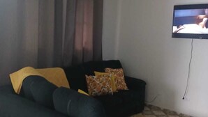Un saló amb un sofà negre, una televisió penjada a la paret i un ventilador de sostre.