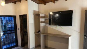 Une pièce avec un téléviseur fixé au mur, un bureau en bois, une porte avec un portail en métal et un sol carrelé.