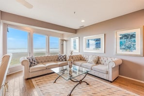 A living room with a sectional sofa, a glass coffee table, and framed artwork on the walls. - Promenade 502 - Top-Floor Oceanfront Penthouse (Seaside)