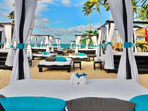 A beachside hotel room with a bed, two pillows, a towel, and a view of the ocean and palm trees. - Luxury 3BR Villa | Private Chef, Golf Cart & Pool | VIP Beach Access (Cabarete)