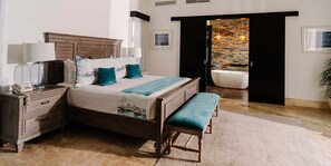A bedroom with a bed, bedside tables, a bench, a bathtub, and a view of a stone wall. - Luxury 3BR Villa | Private Chef, Golf Cart & Pool | VIP Beach Access (Cabarete)