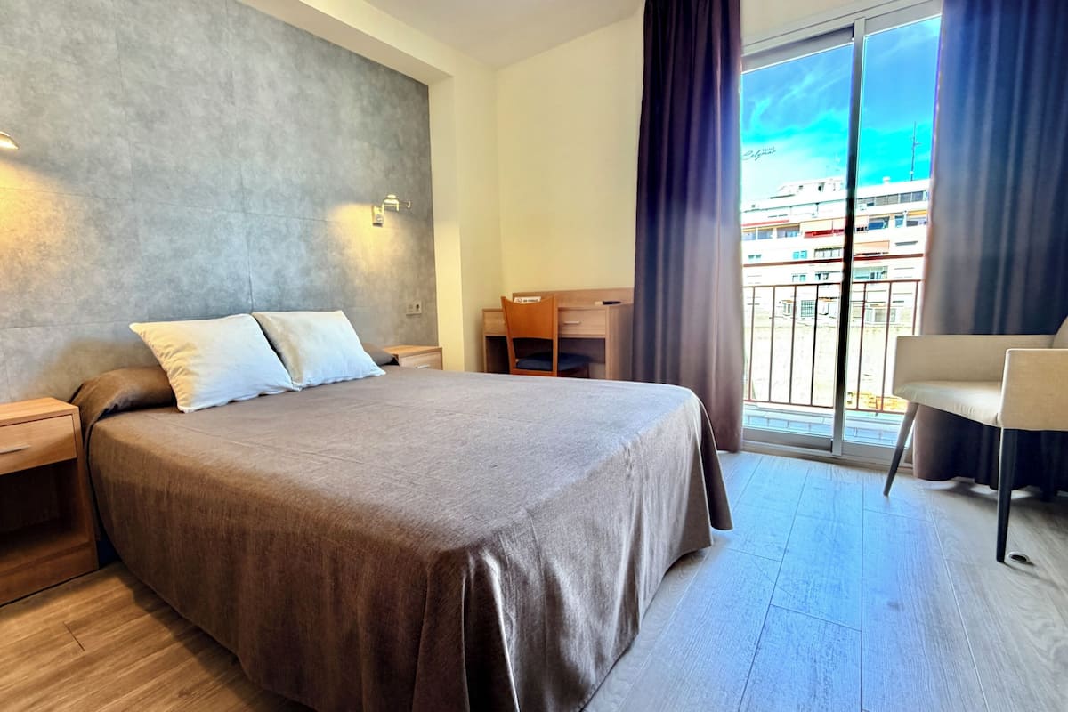 Une chambre d’hôtel avec un grand lit, un bureau, une chaise, un balcon donnant sur les bâtiments et une fenêtre avec des rideaux.